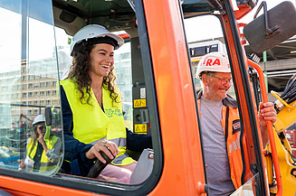 Foto van een medewerker van UZ Gent in een graafmachine, met naast haar een STRABAG-Belgium arbeider tijdens de eerste spadesteek van de nieuwe projecten op UZ Gent.