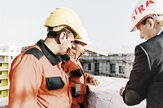 STRABAG werfteam op een STRABAG werf Foto van een STRABAG werfteam in veiligheidskledij met een leidinggevende op een STRABAG werf. Ze hebben allen een helm op. De leidinggevende heeft een kostuumvest aan en een STRABAG helm op.