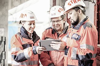 Foto van een team van STRABAG in STRABAG veiligheidskledij op een STRABAG werf. Ze kijken naar een tablet.