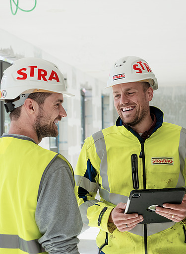 Foto van een team op en werf van STRABAG. Twee manenlijke en één vrouwelijke STRABAG medewerker wandelen op een werf van STRABAG. Ze hebben alle drie STRABAG veiligheidskledij aan. 