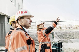 Foto van drie STRABAG medewerkers in oranje STRABAG werfkledij staan op een werf te kijken naar het gebouw in aanbouw. De vrouwelijke medewerker wijst naar boven.           